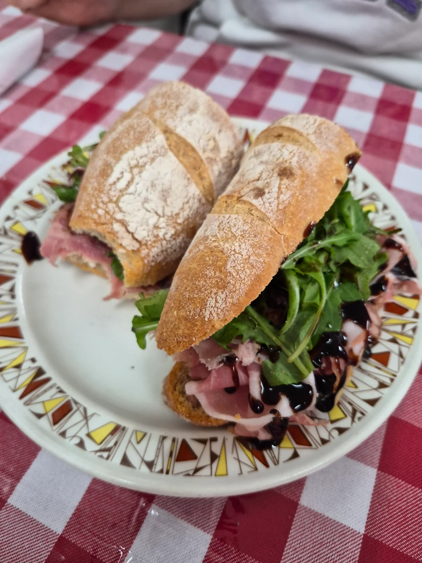 A Big Carm’s sandwich with cured meat, greens and balsamic glaze on a checkered tablecloth